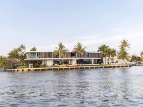 A view of a modern waterfront property surrounded by palm trees, captured from the water. The image lacks clarity, making it difficult to discern details.