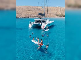 A lively group enjoys a day on the water, paddleboarding and swimming near a catamaran in crystal-clear waters — perfect for summer adventures and family fun.