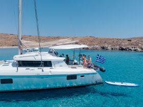 A cheerful group enjoys a sunny day aboard a catamaran in Greece, showcasing a vibrant atmosphere with a paddleboard nearby.