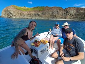 A cheerful group enjoys a day on the water, sharing snacks and laughter aboard their boat, with a scenic rocky backdrop enhancing the experience.