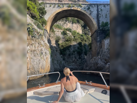 A woman enjoys the stunning view of a historic bridge while relaxing on the deck of a boat, surrounded by dramatic coastal scenery.