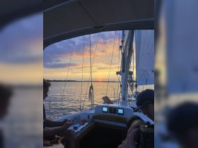 A serene sunset view from a sailboat, with guests enjoying the moment — one playing guitar while another captures the scene, perfect for a relaxing evening on the water.