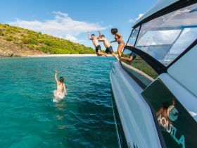 A lively group enjoys a fun day on the water, with friends jumping off the boat into the turquoise sea — perfect for adventurous beach outings.