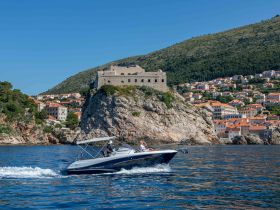 A sleek boat cruises along the stunning coastline, with a historic fortress perched on a rocky outcrop in the background, showcasing the beauty of the region.