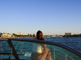 A woman enjoys a serene sunset view from the deck of a boat, overlooking a picturesque waterfront — a perfect moment for relaxation and scenic beauty.