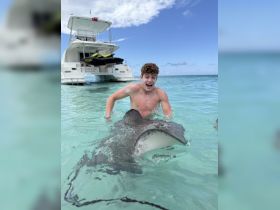 A young man joyfully interacts with a stingray in shallow waters, showcasing an exciting wildlife encounter during a boating adventure.