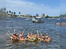 A lively group of friends enjoys a fun day in the water, celebrating with drinks and pool noodles amidst a backdrop of boats — perfect for summer parties and gatherings.