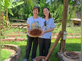Two women smile while holding a decorative plate in a lush outdoor setting, showcasing a moment of cultural experience.