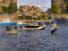 A sleek boat navigates the water with passengers enjoying the scenic view of unique architectural buildings in the background, perfect for urban boating experiences.