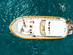 A lively group enjoys a sunny day on a boat, with some relaxing on deck and others swimming in the crystal-clear water — perfect for a fun-filled day on the water.