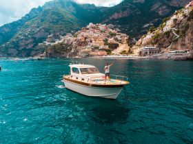 A joyful individual waves from the deck of a boat, surrounded by the stunning Amalfi Coast, showcasing the vibrant scenery and inviting waters perfect for exploration.