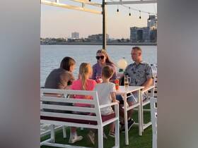A family enjoys drinks and conversation at a waterfront dining area, capturing the essence of a relaxing evening by the water.