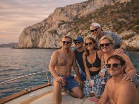 A joyful group of friends enjoys a sunset boat trip, celebrating together with drinks and laughter against a stunning coastal backdrop.