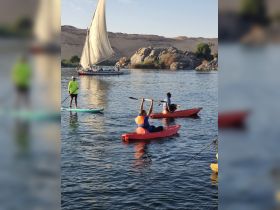 A lively group enjoys kayaking on the river, with a traditional sailboat in the background, showcasing the perfect blend of adventure and scenic beauty.