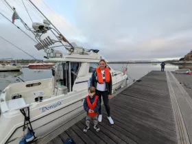 A woman and a child pose in life jackets next to the boat 'Donzela' at the dock, ready for a fun day on the water.