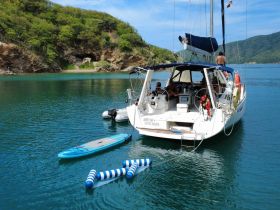 A scenic view of a sailboat anchored in tranquil waters, with guests enjoying the sun and a paddleboard nearby — perfect for a relaxing day on the water.