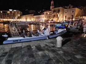 A sleek boat is docked at a lively harbor at night, illuminated by the lights of nearby buildings, showcasing a vibrant waterfront atmosphere.