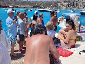 A lively group enjoys a fun day on the water, celebrating with drinks and laughter aboard a boat, surrounded by stunning blue waters and rocky scenery.