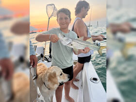 A young boy proudly holds up his catch while fishing at sunset, accompanied by a friendly dog — a perfect moment of family fun on the water.