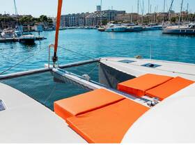 A vibrant view of the lounge area on a catamaran, featuring bright orange cushions against a backdrop of a serene marina — perfect for relaxing on the water.