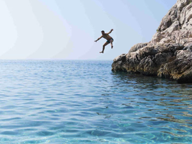 A person joyfully leaps off a rocky ledge into the crystal-clear ocean, capturing the thrill of summer adventures and water activities.