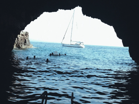 A group of swimmers enjoys the refreshing waters inside a cave, with a sailboat anchored just outside — a perfect blend of adventure and scenic beauty.