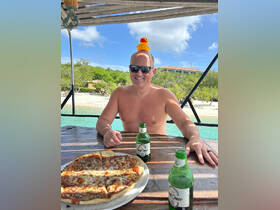 A man enjoys a relaxing moment with pizza and beer on a boat, complete with a playful rubber duck on his head — a perfect snapshot of fun in the sun.