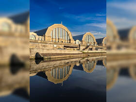A scenic view of industrial buildings reflected in calm waters, showcasing architectural design against a clear sky.