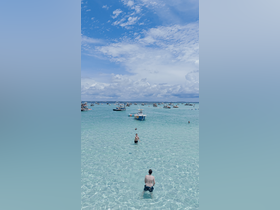 Two friends enjoy a playful moment in the shallow waters, tossing a ball while surrounded by boats — a perfect day for beach fun and relaxation.
