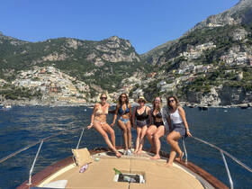 A group of friends poses on the bow of a boat, enjoying a sunny day with a stunning coastal backdrop — perfect for memorable outings along the Amalfi Coast.