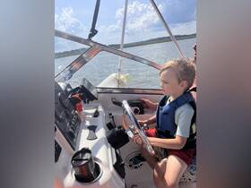 A young child takes the helm of a boat while an adult assists, showcasing a fun family boating experience on the water.