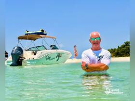 A man enjoys a sunny day in shallow waters near a boat, with a beach backdrop and another person in the distance — a perfect scene for summer adventures.