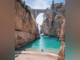 A stunning view of a couple standing in shallow water at a picturesque beach, framed by dramatic cliffs and a boat anchored nearby — a perfect representation of coastal adventures.