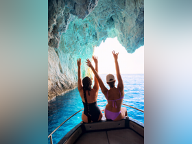 Two friends celebrate their adventure aboard a boat, raising their hands in excitement while exploring a stunning cave along the coastline — a perfect moment for unforgettable memories on the water.