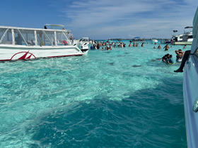 A lively scene with groups of people enjoying the clear waters, perfect for swimming and fun in the sun. The boats provide a backdrop to this vibrant beach experience.