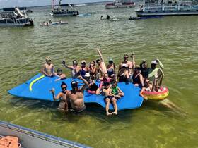 A lively group enjoys a fun day on a floating mat in shallow waters, celebrating with drinks and laughter — the perfect summer party atmosphere.