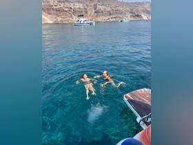 Two friends enjoy a refreshing swim in the crystal-clear waters, with a scenic backdrop of cliffs and a sailboat nearby — perfect for summer adventures.