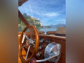 A close-up view of the helm inside a classic wooden boat, showcasing the steering wheel and control panel, with a scenic backdrop of a lakeside villa.