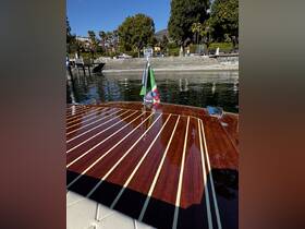 A close-up view of a beautifully crafted boat deck featuring polished wood and a flag, set against a scenic backdrop of trees and a dock.