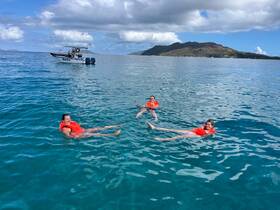 A group of friends enjoys a refreshing swim in crystal-clear waters while wearing life jackets, with a boat anchored nearby and a scenic island backdrop.