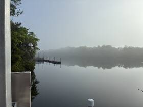 A foggy morning scene over a calm body of water, featuring a distant dock and reflections, creating a serene atmosphere.