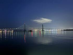 A nighttime view of a bridge reflecting on the water, creating a serene atmosphere. The image captures the beauty of the structure illuminated against the dark sky.