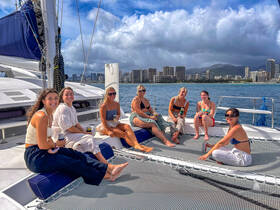 A joyful group of friends enjoys drinks aboard a catamaran, with stunning views of the city skyline in the background — perfect for a fun day on the water.