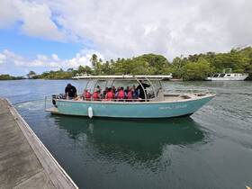 A group of divers prepares to embark on an adventure aboard the Tropical Diver boat, surrounded by lush greenery and calm waters, perfect for exploring underwater wonders.