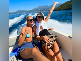 Two friends enjoy a sunny day on a boat, celebrating with smiles and thumbs up against a stunning backdrop of mountains and water — perfect for memorable outings.