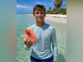 A young boy proudly holds a large conch shell while standing in shallow water on a beautiful beach, showcasing a delightful moment of beachcombing and exploration.