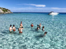 A lively group enjoys a refreshing swim in crystal-clear waters, with a boat anchored nearby, perfect for a fun day at the beach.