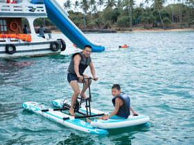 A father and son enjoy a fun day on the water, paddleboarding together near a boat with a water slide, capturing the essence of family adventure and water activities.