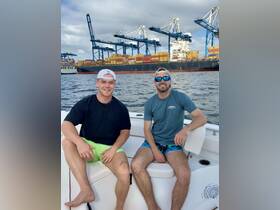 Two friends enjoy a day on the water, relaxing on a boat with a bustling harbor backdrop filled with cargo ships and cranes.
