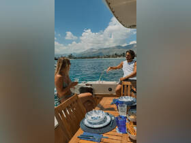 A couple enjoys drinks and a beautifully set dining area on a boat, surrounded by stunning coastal scenery — perfect for a relaxing day on the water.